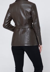 Giacca di pelle marrone con design aderente, vita con cintura e polsini con cerniera. Tessuto liscio e cuciture strutturate. Vista posteriore mostrata.
