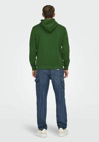 Felpa felpata verde con cappuccio regolabile, abbinata a pantaloni cargo blu con tasche laterali, indossati con sneakers bianche.
