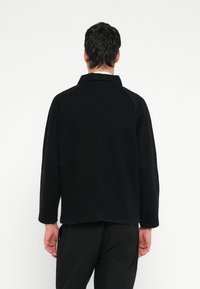 Čierna rifľová bunda s golierom, raglánovými rukávmi a rovnakým strihom. Charakterizuje ju prešívanie a hladká textúra.