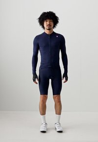 Navy Radoutfit mit langen Ärmeln, einem Reißverschluss vorne und enganliegenden Shorts. Enthält schwarze fingerlose Handschuhe und weiße Fahrradschuhe.
