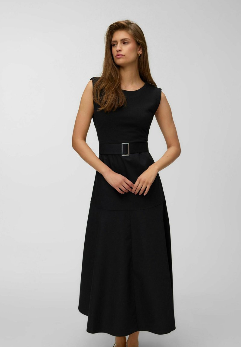 Femme aux longs cheveux bruns portant une robe noire sans manches avec une ceinture, debout les mains jointes, regardant sur le côté sur un fond uni.