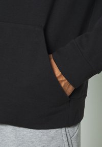 Sudadera negra con un bolsillo frontal y puños acanalados, llevada sobre pantalones de chándal gris claro. El tejido parece suave y texturizado.