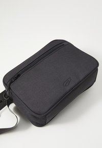 Schwarze rechteckige Tasche aus strukturiertem Stoff, mit einem Reißverschluss oben und einem verstellbaren Riemen. Minimalistisches Design mit Logo-Detail.