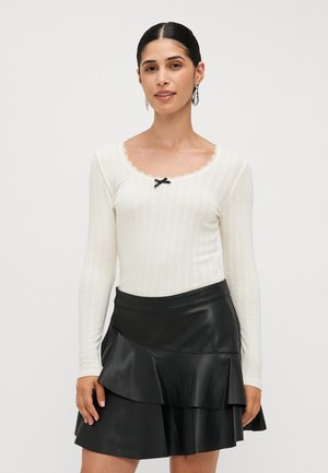 White ribbed long-sleeve top with a scalloped neckline and a black bow accent, paired with a black faux leather flared mini skirt.