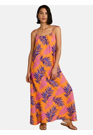 Vrouw draagt een oranje maxi-jurk met paarse bladmotieven en dunne schouderbandjes, gecombineerd met beige platte sandalen op een witte achtergrond.