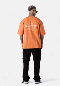 T-shirt en coton orange avec un texte graphique au dos, associée à un pantalon cargo noir. Design décontracté avec des manches courtes et une coupe ample.