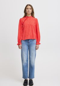 Röd långärmad blus med vita prickar, volangärmar och knapprad, kombinerad med ljusblå raka jeans och svarta platåskor.