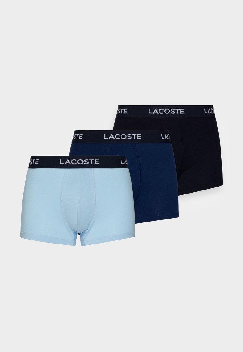 Tre par kalsonger i ljusblått, marinblått och svart. Varje par har en kontrasterande svart midjeband med "Lacoste"-logotyp. Slät tygtextur.