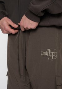Bruine oversized hoodie met ribgebreide manchetten, gecombineerd met lichte bruine cargobroek met verstelbare trekkoorden en een prominent logo.