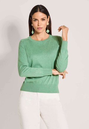 Frau mit glattem dunklem Haar, die einen mintgrünen Pullover, weiße Hosen, grüne geometrische Ohrringe und goldene Ringe trägt und mit der Hand nahe am Gesicht posiert.