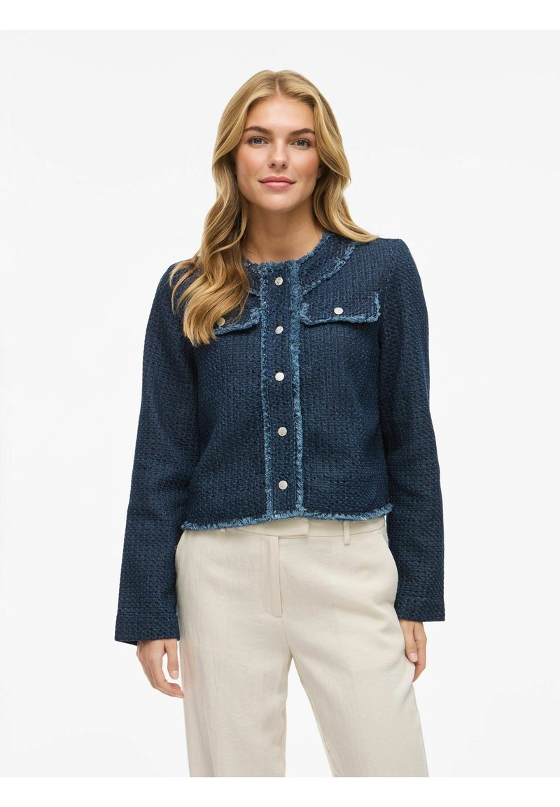 Tamno plava tkanina jakna s dva prednja džepa, izgužvanih rubova, okruglim izrezom i srebrnim gumbima, uparenih sa svijetlim hlačama.