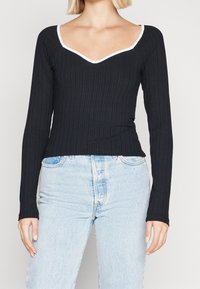 Zwarte geribbelde longsleeve top met een V-hals en witte afwerking, gecombineerd met lichtblauwe high-waisted jeans. De textuur is glad en aansluitend.