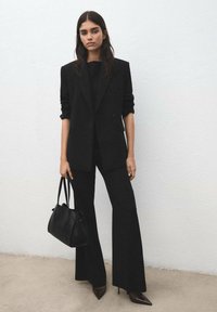 Czarna, dopasowana marynarka na top dopasowany i rozkloszowane spodnie. Modelka trzyma czarną torebkę i nosi szpilki z zamkniętym, szpiczastym noskiem. Gładki, minimalistyczny design.