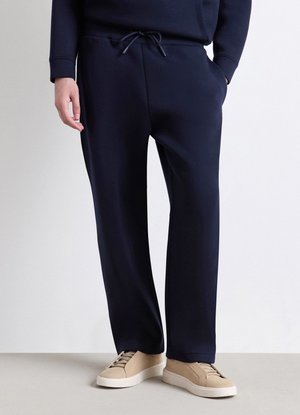 Uomo che indossa pantaloni blu navy con coulisse e sneakers beige, in piedi davanti a uno sfondo chiaro e semplice con una mano in tasca.
