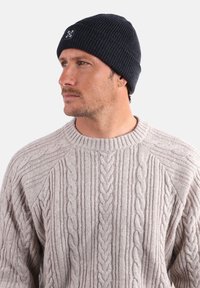 Homme regardant à gauche portant un bonnet en tricot noir avec un logo et un épais pull en tricot beige.