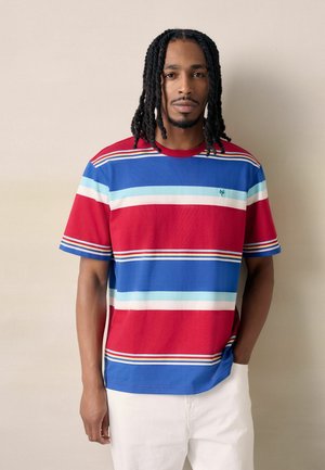 Man with dreadlocks wearing a red, blue, white striped short-sleeve shirt and white pants, standing against a beige background.