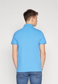 Polo shirt bleu clair avec un col et des manches courtes, présentant un tissu texturé et des détails de couture. Porté avec un jean bleu foncé.