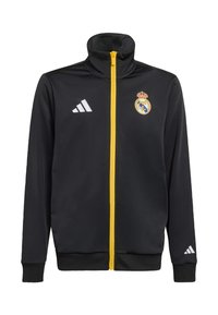 Musta urheilutakki, jossa on korkea kaulus, keltainen vetoketju ja valkoinen Adidas-logo. Rintakehässä Real Madridin tunnus.
