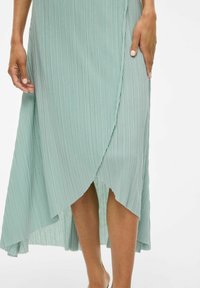 Light green pleated maxi dress with a wrap design, featuring a high-low hem and a soft, textured fabric. Exposed hand resting on the side.