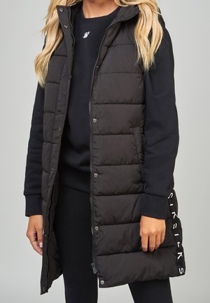 Long gilet noir matelassé sans manches, porté ouvert sur un sweat-shirt noir et un pantalon, avec les cheveux blonds visibles aux épaules.