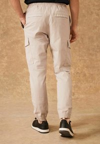 Pantaloni cargo beige con polsini elastici, dotati di tasche laterali e tasche posteriori per la funzionalità. Indossati con sneakers nere su uno sfondo testurizzato.