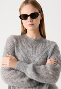 Grauer, halb transparenter Pullover mit geripptem Textur und breiten, weichen Ärmeln. Das Model trägt schwarze ovale Sonnenbrillen. Die Hände sind an den Ellbogen verschränkt.