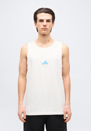 Joven de pie, vistiendo una camiseta blanca sin mangas con un pequeño logo azul en el pecho y pantalones negros, sobre un fondo liso.