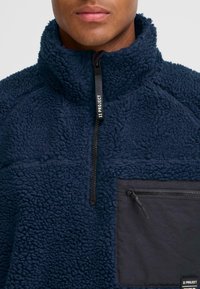 Marineblauer Fleece-Pullover mit hohem Kragen und Viertelreißverschluss vorne. Verfügt über eine kontrastierende schwarze Reißverschlusstasche auf der Brust und ein strukturiertes Material.