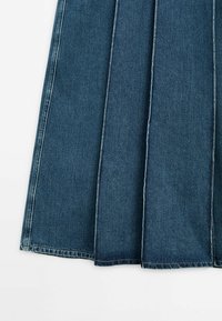Tissu en denim bleu avec de larges plis verticaux cousus, formant une partie d'une jupe évasée sur fond blanc.