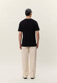 Melns kokvilnas T-krekls ar īsām piedurknēm un apaļu kakla izgriezumu, valkāts kopā ar krēmbaltām platām biksēm, skatīts no aizmugures pret neitrālu fonu.