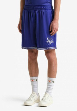 Uomo con pantaloncini sportivi blu con logo ricamato vicino all'orlo, calzini bianchi con dettagli arancioni e verdi, e scarpe da ginnastica bianche, in piedi.