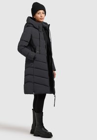 Czarna pikowana kurtka z kapturem, z zapięciem na zamek, bocznymi kieszeniami i geometrycznym wzorem, w połączeniu z czarnymi butami na sznurówki.