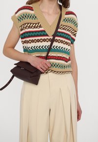 Chaleco de punto con cuello en V, con rayas en beige, verde, rojo y marrón. Combinado con pantalones plisados color crema y un pequeño bolso cruzado marrón.