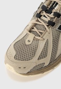 Chaussure de sport beige avec tissu en maille, semelle en caoutchouc et lacets noirs. Dispose d'un embout moulé et d'un logo 'N' bien visible sur le côté.