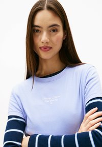 Camicetta lilla a maniche corte con testo "Tommy Jeans New York City" in bianco. Maniche lunghe a righe blu navy sotto, in morbido cotone.