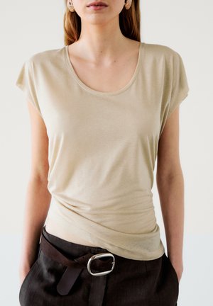 Woman wearing beige short-sleeve top tucked into dark pants with a large silver buckle belt, standing against a light background.