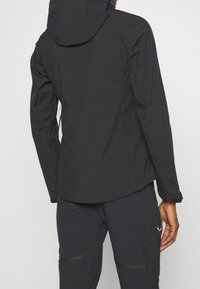 Veste extérieure noire avec capuche, fabriquée en tissu lisse et résistant à l'eau ; dotée d'un ourlet arrière plus long et de poignets élastiques.