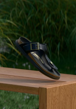 Sandale noire Birkenstock avec boucle reposant sur une table en bois à l'extérieur avec des plantes vertes floues en arrière-plan.