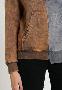 Kapuzenpullover mit einem Farbverlauf in Braun- und Blautönen, ausgestattet mit einer Kängurutasche vorne und gerippten Bündchen. Weiche Textur.