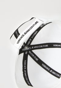 White bra featuring black Versace Jeans Couture straps with logo print, smooth fabric, and rounded cups with a subtle sheen.