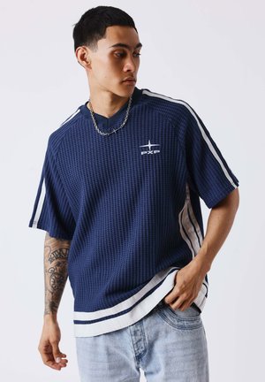 Joven con cabello corto y negro, vestido con una camisa azul texturizada con rayas blancas y vaqueros azul claro, mirando hacia su izquierda.