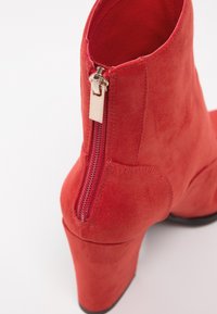 Bottine en daim rouge avec un talon bloc haut, dotée d'une fermeture éclair sur le côté et d'une texture lisse. L'intérieur est également doublé en rouge.