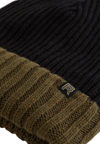 Stickad mössa i svart och olivgrönt. Har ribbat mönster och en vikbar kant. Svart etikett med vitt logo på sidan.