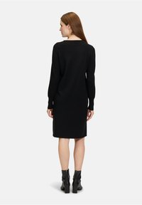 Robe noire à manches longues avec une silhouette droite, un col rond et une longueur juste au-dessus du genou, associée à des bottines noires.