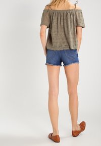 Blusa verde oliva sin hombros con patrón texturizado, combinada con pantalones cortos de mezclilla azules y zapatos planos marrones, vista desde la espalda.