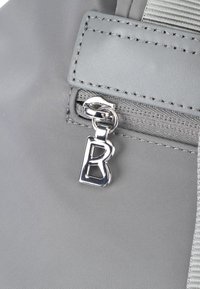 Bolsa de tela gris con cremallera, con un tirador de hardware plateado que tiene un charm de "B". Textura suave, detalles cosidos y una correa duradera.