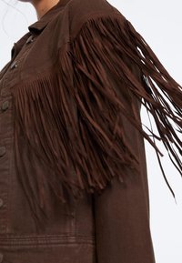 Bruine spijkerjack met franjes op de mouwen, een gestructureerde afwerking en metalen knoopsluitingen.