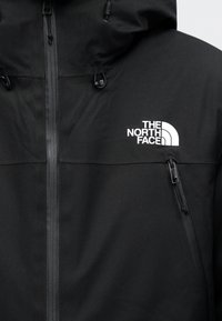 Veste noire à capuche avec fermeture éclair à l'avant, poche poitrine zippée et logo blanc "The North Face" sur la poitrine gauche.
