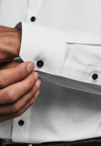 Camisa blanca de vestir con tejido texturizado, acentos de botones negros en los puños y un primer plano de una mano ajustando el puño.