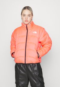 Giacca a vento arancione brillante con design trapuntato, chiusura con zip, colletto alto e dettaglio del logo; abbinata a pantaloni corti neri di pelle.